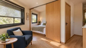 Interior de tiny house com poltrona azul-marinho, cama parcialmente protegida por painel deslizante de madeira e janela com persiana regulável.