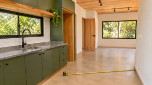 Interior de Tiny House em fase de acabamento, com cozinha linear verde-musgo, bancada cinza e trena medindo o espaço de circulação.